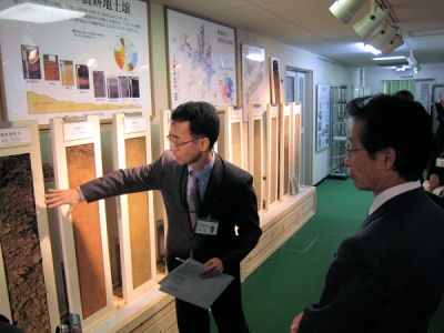 写真２ 土壌モノリス（土壌断面標本）の前で日本の土壌についての説明を聞かれる佐々木政務官 （説明者 谷山研究コーディネータ）